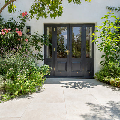 A stylish black double door with glass panels, framed by lush greenery and vibrant flowers, opens onto a sunlit Hambleton Ivory patio .