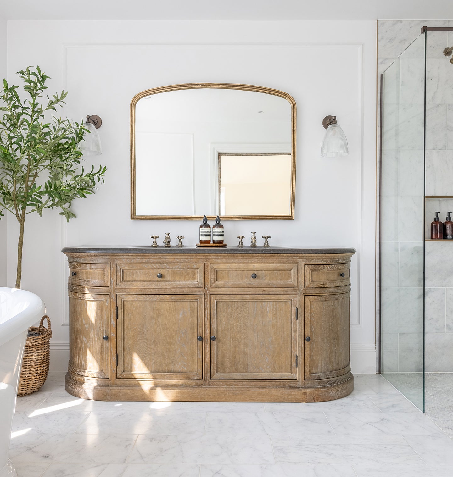 Elegant bathroom with Carrara Bianco marble tiles with a rustic wooden vanity, large mirror, marble shower, and bright window. A potted plant and basket add natural elements. Serene ambiance.