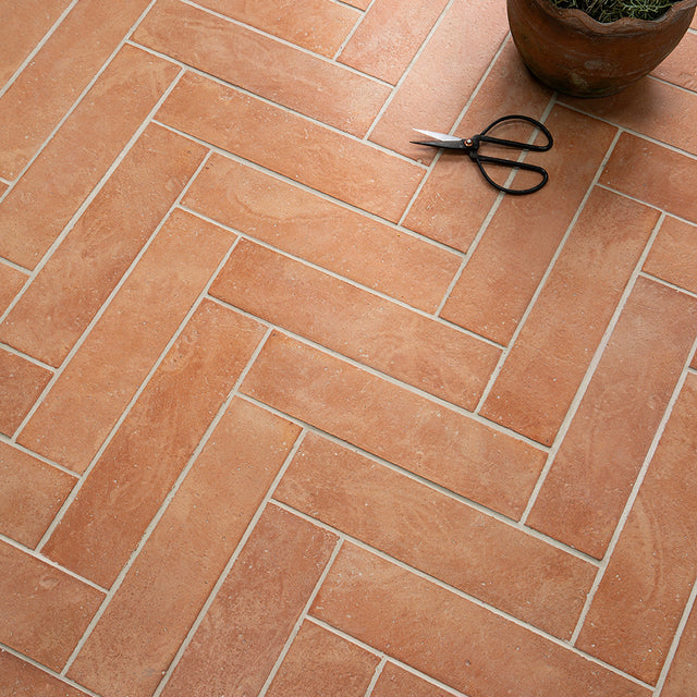Terracotta tiles arranged in a herringbone pattern with a pair of scissors and a plant pot on a similar surface.