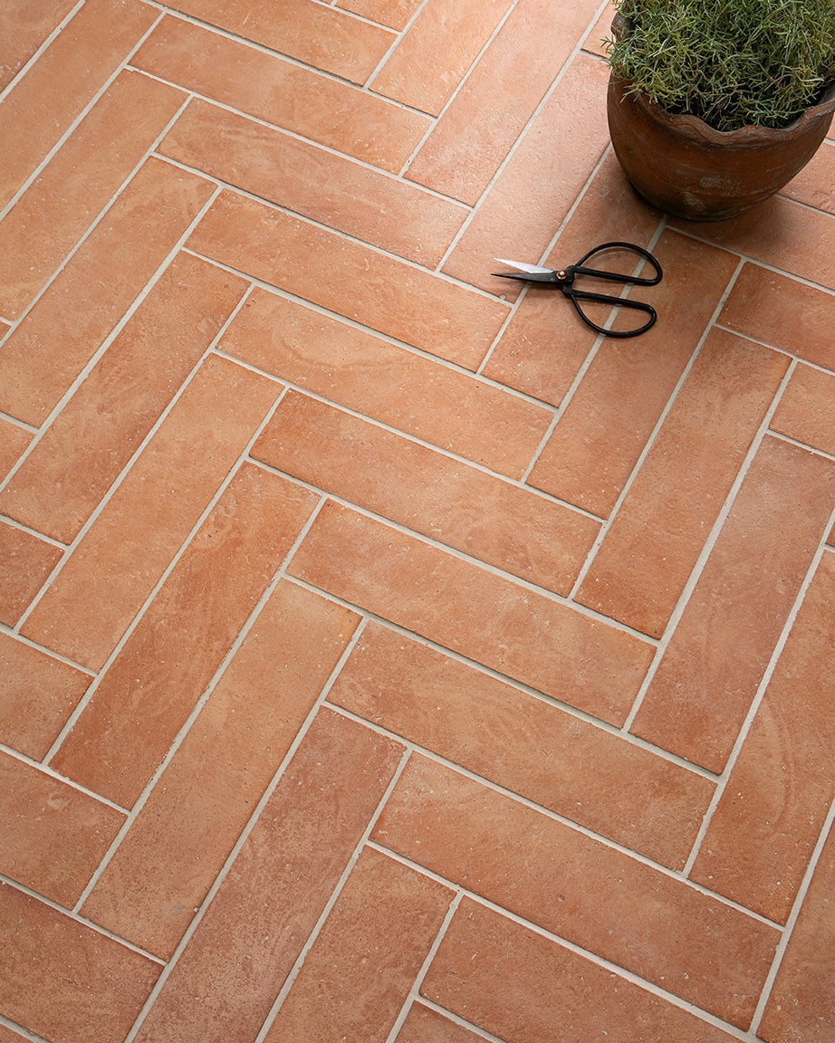 Terracotta tiles arranged in a herringbone pattern with a pair of scissors and a plant pot on a similar surface.